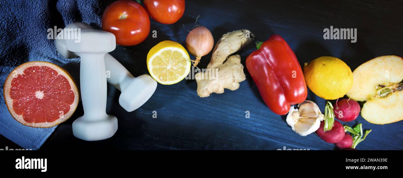 Dumbbells, vegetables and fruits on a dark blue wooden background, wholesome lifestyle with sport and healthy food, panoramic format, top view from ab Stock Photo