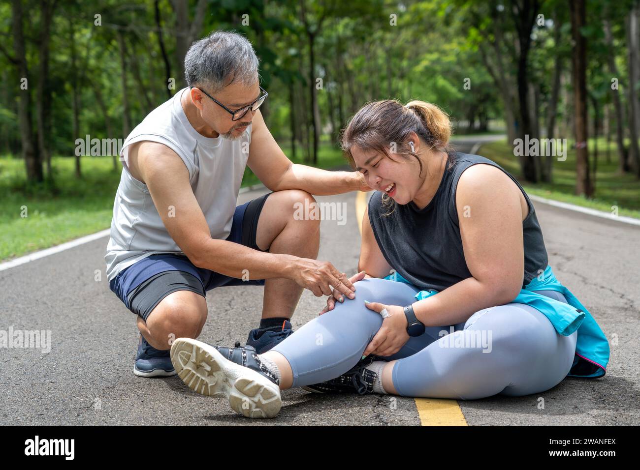 Overweight young woman with knee pain sitting on the running track at local park with both hands grabbing on her trouble knee while a middle age man k Stock Photo