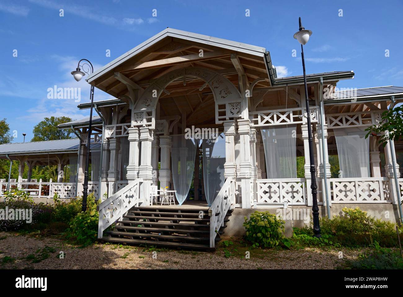An elegant wooden veranda with white columns and stairs, surrounded by plants under a blue sky, Old railway station Bialowieza Towarowa, built for Rus Stock Photo