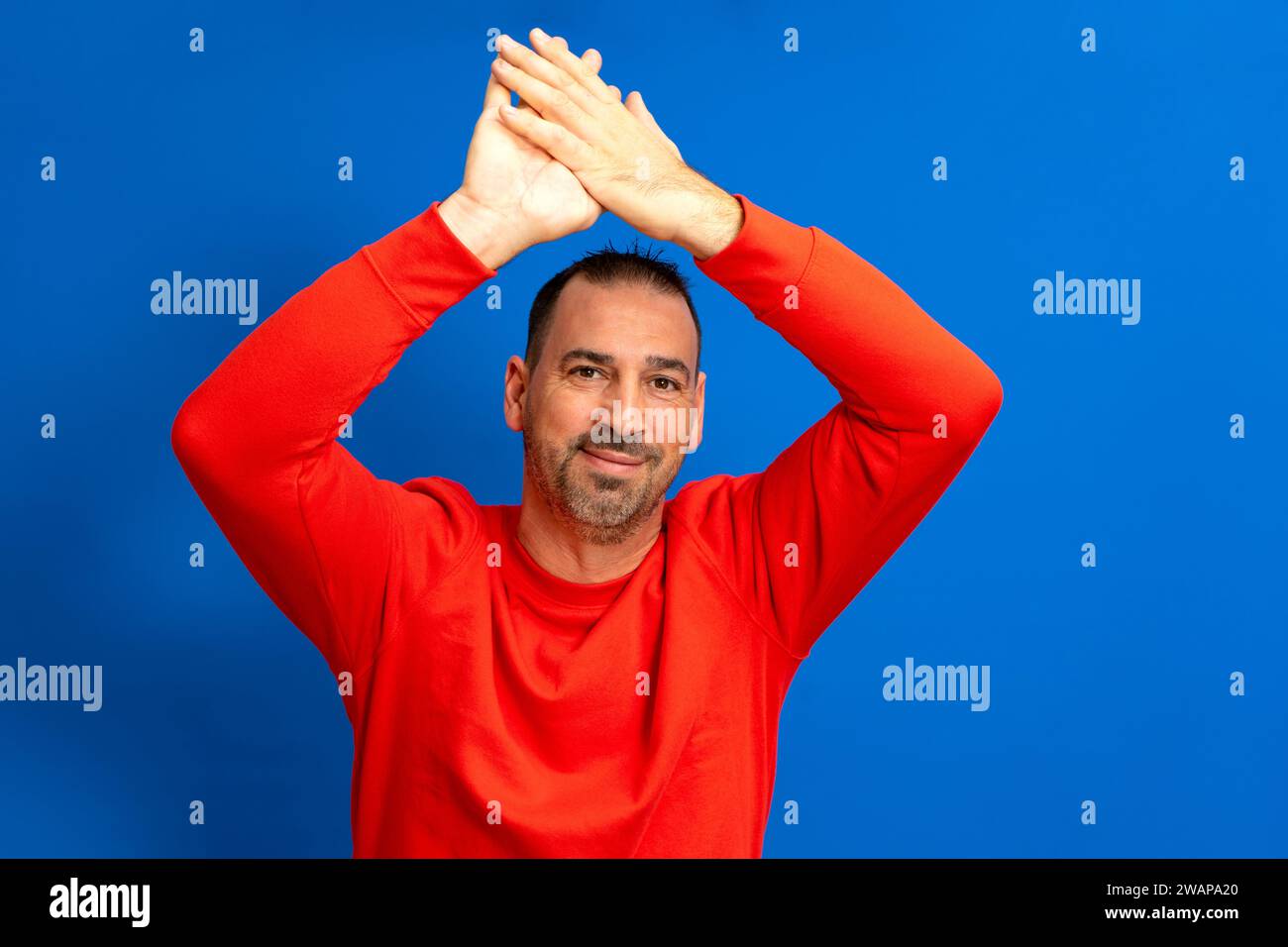 Hispanic man with beard in his 40s clapping with his hands above his head isolated on blue studio background, he is excited about the great spectacle Stock Photo