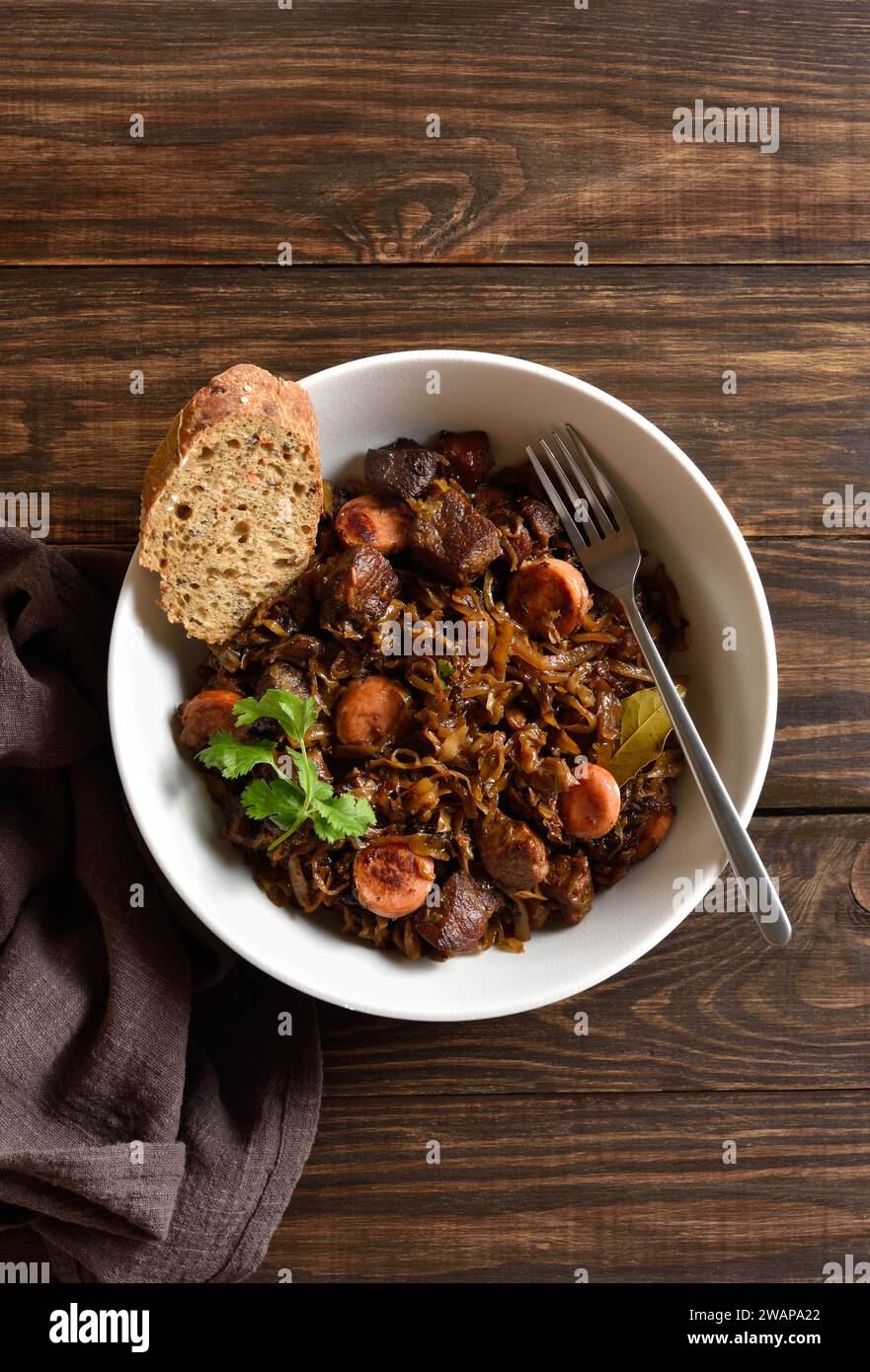 Stewed cabbage (polish bigos) with sauerkraut, mushrooms, smoked meats and spices in bowl over wooden background. Top view, flat lay Stock Photo