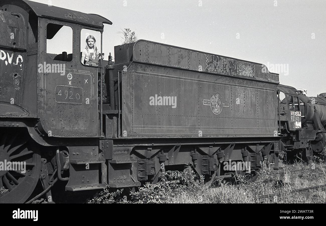 1975, historical, a young girl standing in the driver's cab of an old steam locomotive, no, 4920 at the Woodham Brothers scrapyard at Barry Docks, South Wales, commonly known as Barry Scrapyard. Behind the loco's coal wagon, steam locomotive 92240, with the marking Sold written on the side. The Woodham Brothers, scrap metal merchants established in 1892, brought the old Briitsh Railways steam locomotives and then either sold them on or scrapped them. It is estimated that over 200 steam locomotives were rescued for the railway preservation movement. Stock Photo