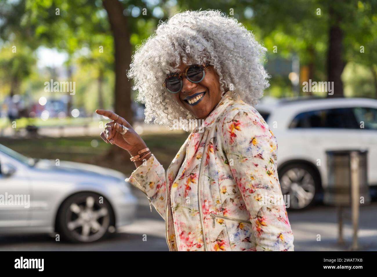 Self-confident, black lady with hoary afro hair and sunglasses smiling while walking in a sunny city center. Concept: pro-aging, happiness Stock Photo