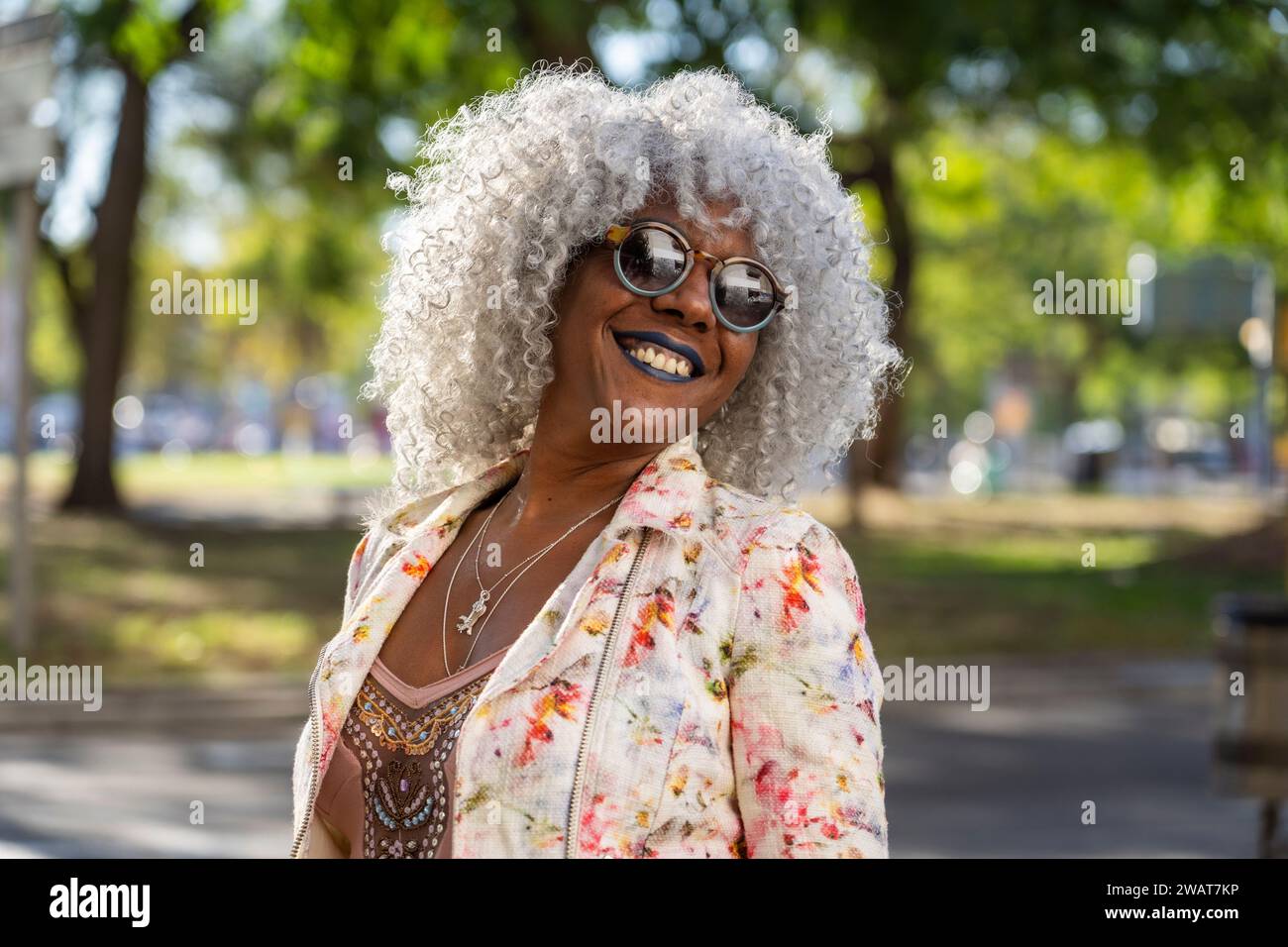 Self-confident, black lady with hoary afro hair and sunglasses smiling while walking in a sunny city center. Concept: pro-aging, happiness Stock Photo