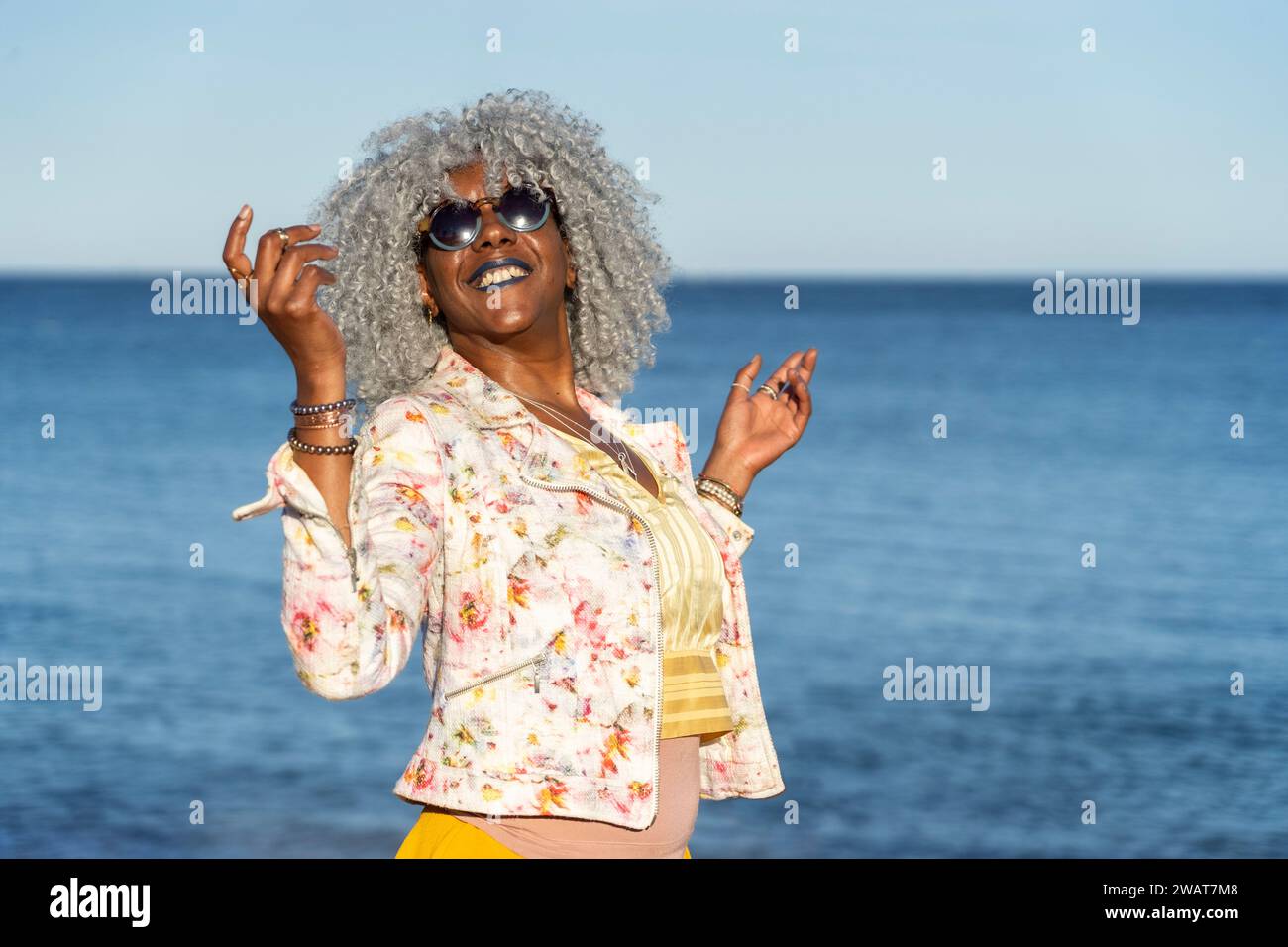Self-confident, black lady with hoary afro hair and sunglasses smiling while walking on a sunny beach. Concept: pro-aging, vacations Stock Photo