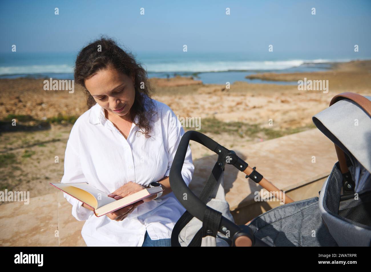 Happy young African American woman, mother pushing baby stroller, reading book, sitting relaxed on the parapet on Atlantic beach. Maternity leave. Peo Stock Photo