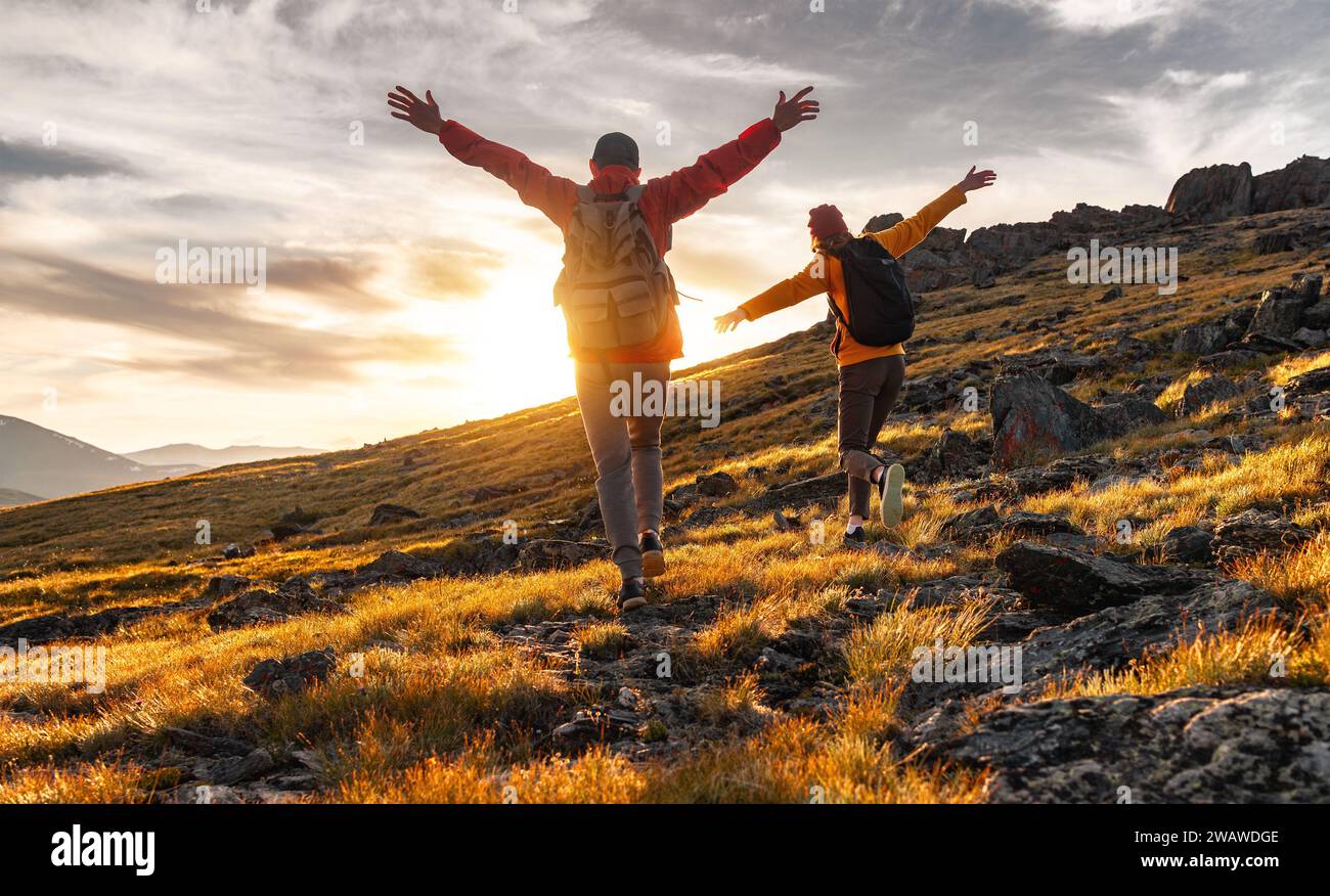 Couple hikers are walking together in sunset mountains. Two young tourists with backpacks are standing with open arms and enjoys sunset Stock Photo