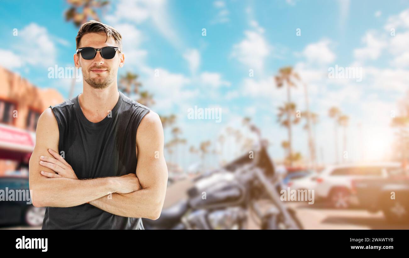Portrait of American young handsome man chopper biker rider  in sleeveless shirt with sunglasses summer coast beach background standing smiling. Stock Photo