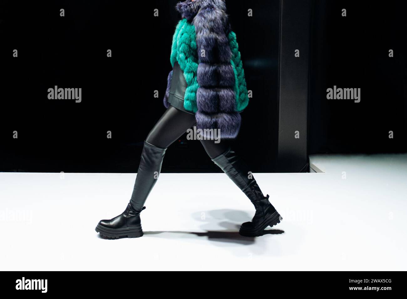 Female figure dressed in a fur coat and black leather boots walking on black background. Stock Photo
