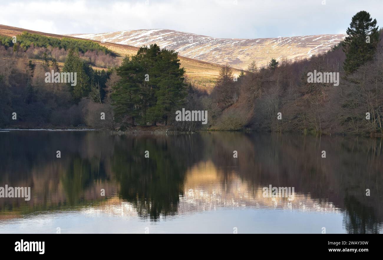 Trees, snow-dusted mountains and reflections at Glenogil reservoir on a dry January day in Angus, Scotland. Stock Photo