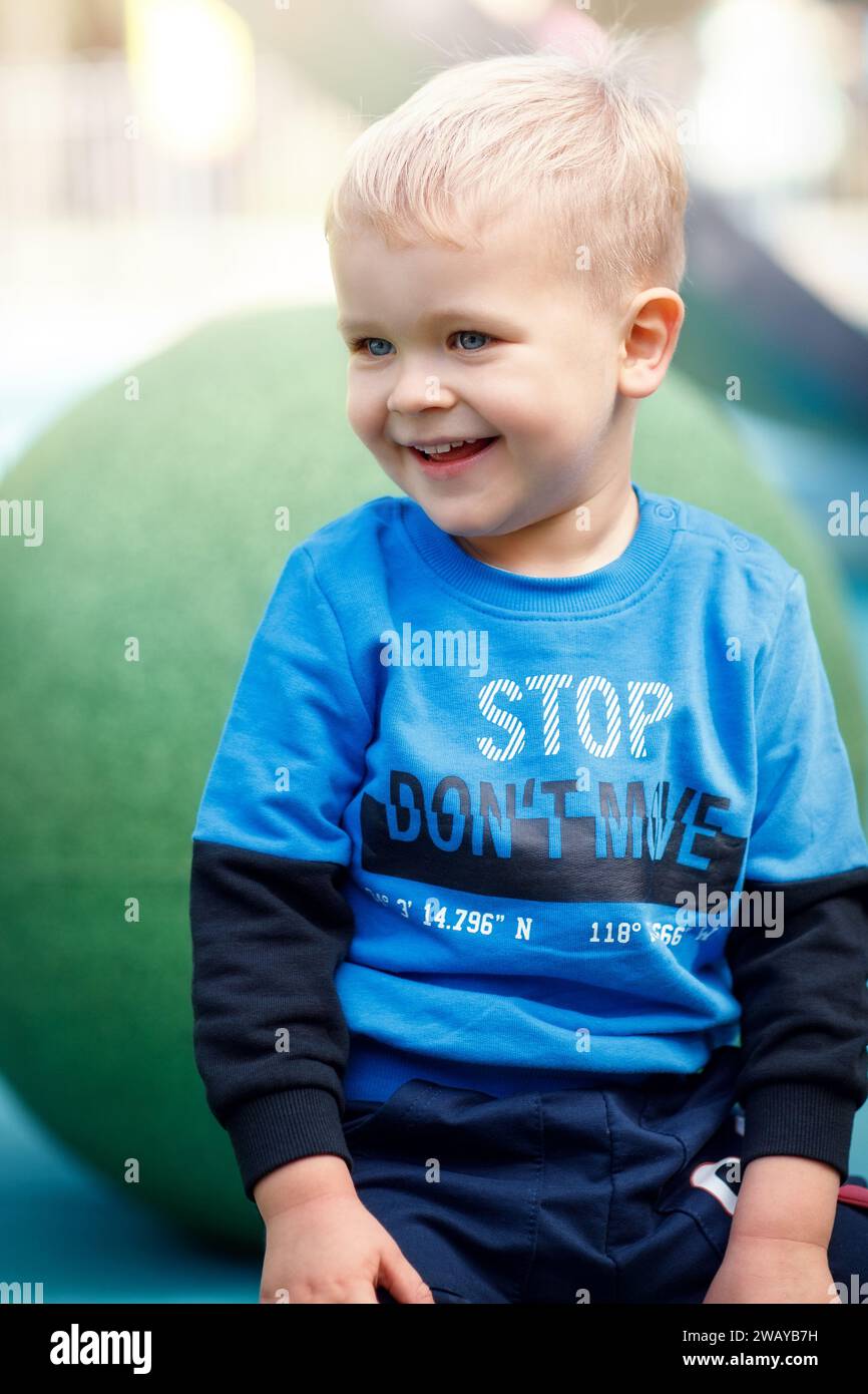 Portrait of a happy and smiling blond little boy sitting on a green ball in the playground. Stock Photo
