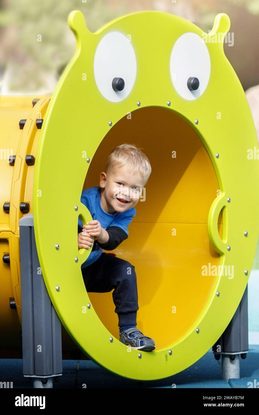 Cheerful smiling boy on sunny summer day in outdoor playground. The child is playing hide-and-seek, he skillfully hides in the yellow obstacle tube re Stock Photo