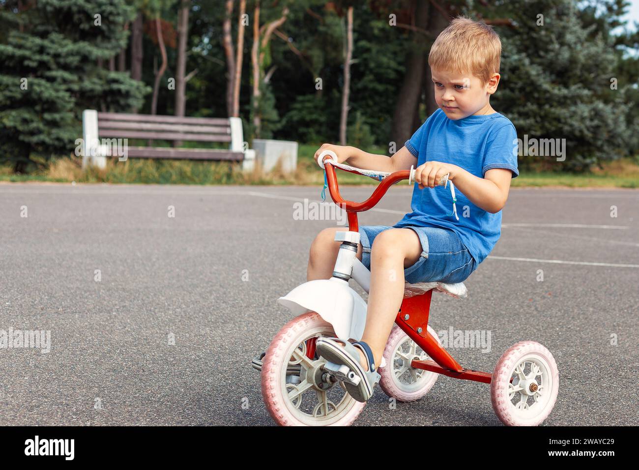 A little boy in blue clothes, very focused rides an red tricycle in city park. Stock Photo