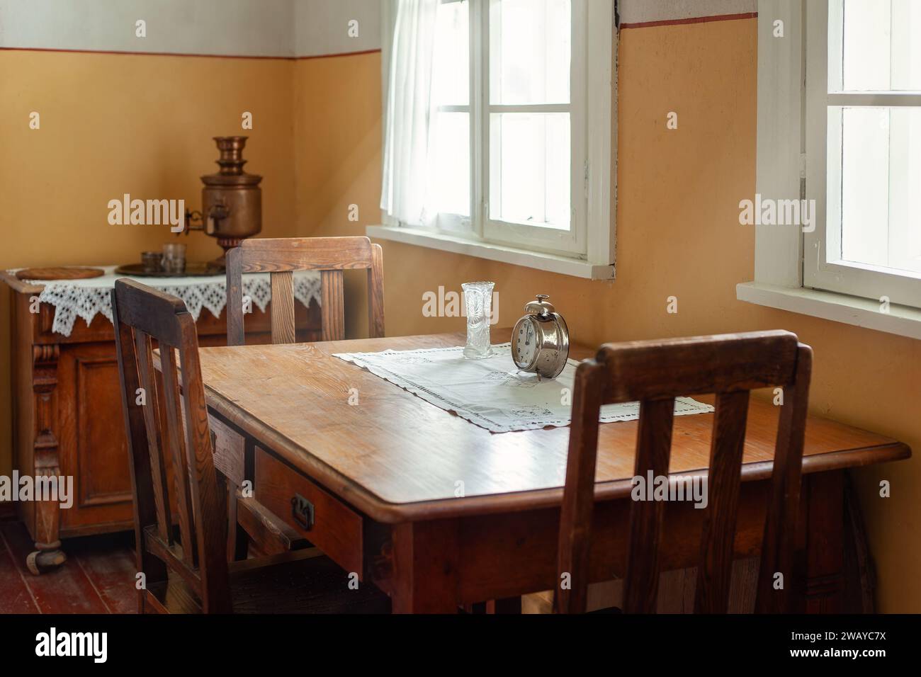 An antique wooden table and three chairs by the window, daylight streaming in to illuminate a baptismal vase and a large antique clock on the table. Stock Photo