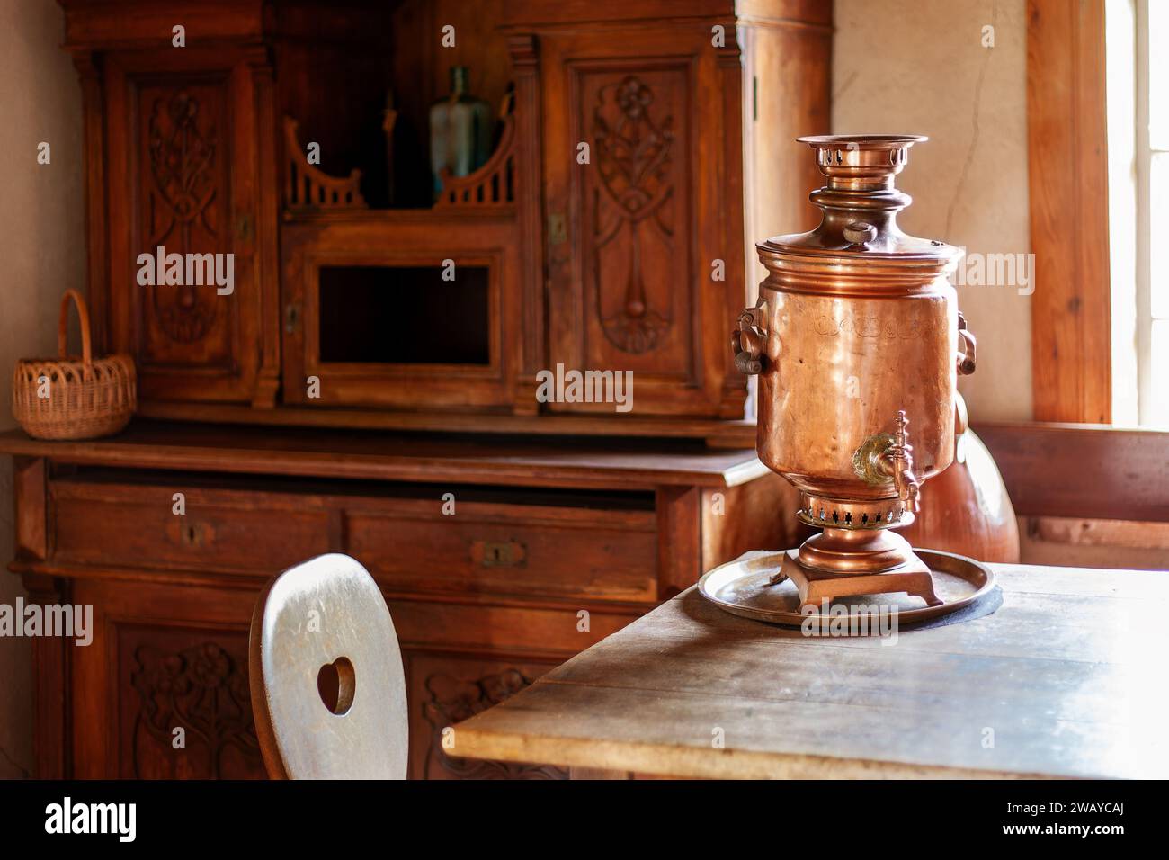 Interior of an ancient guest room. In the room there is a beautiful carved wood cabinet for dishes, a sunlit table and a copper Russian kettle for tea Stock Photo