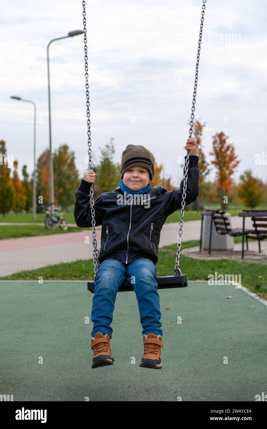 Smiling cute boy in black leather jacket and brown hat swings on swing in city park. Stock Photo