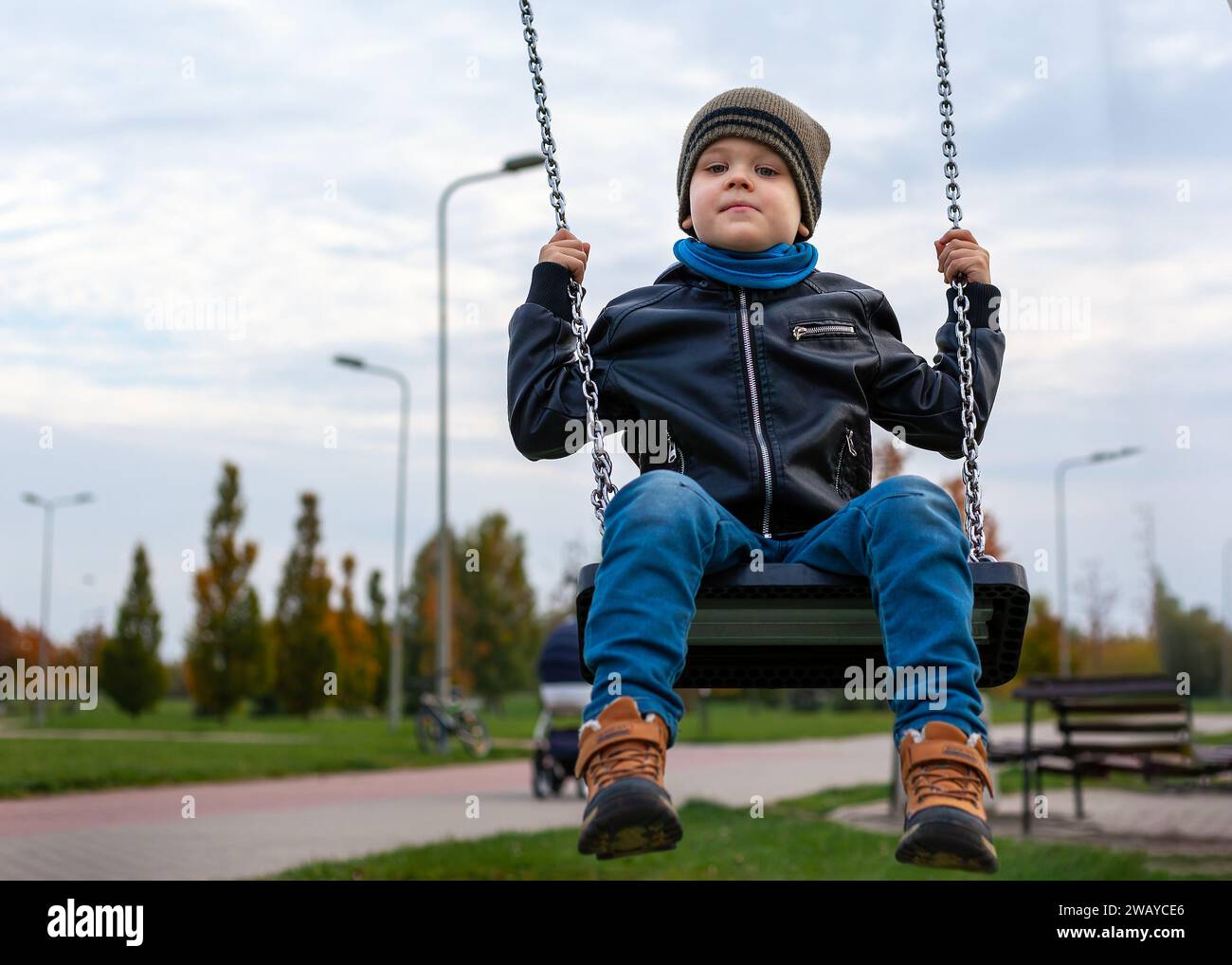 A little boy swings in a city park on a swing, he holds tightly to the metal chains. Stock Photo