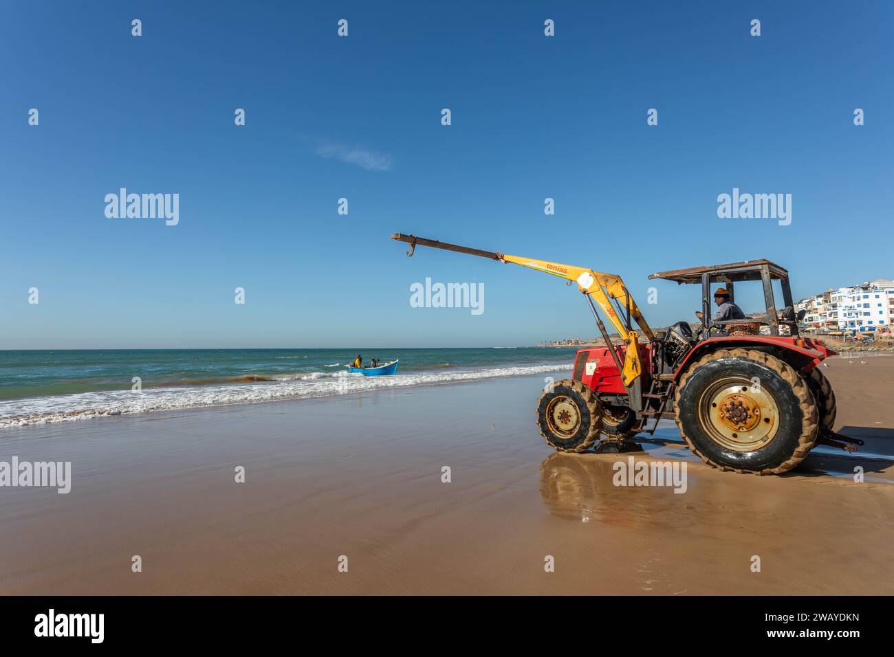 A tractor is on the beach, ready to lift a small, blue, wooden fishing boat from the water after a fishing trip. Taghazout, Morocco, North Africa. Stock Photo