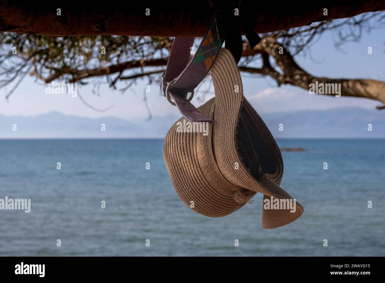 Ladies straw hat hanging on a tree, in an early evening sunlight. Blurry background created by water of Ionian sea and albanian mountains. Paralia Agi Stock Photo