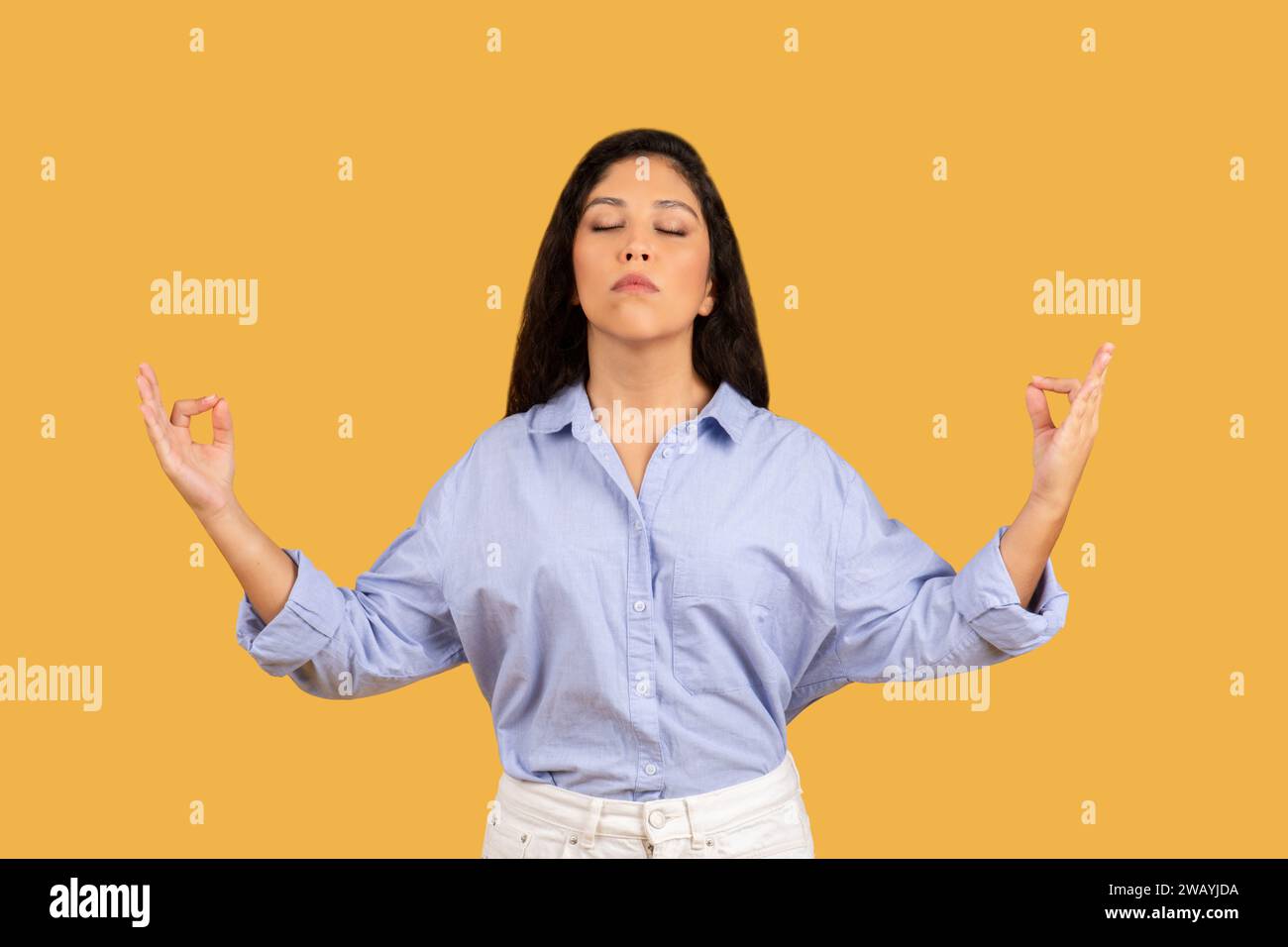 A serene young woman with closed eyes in a light blue shirt practices meditation with raised hands Stock Photo
