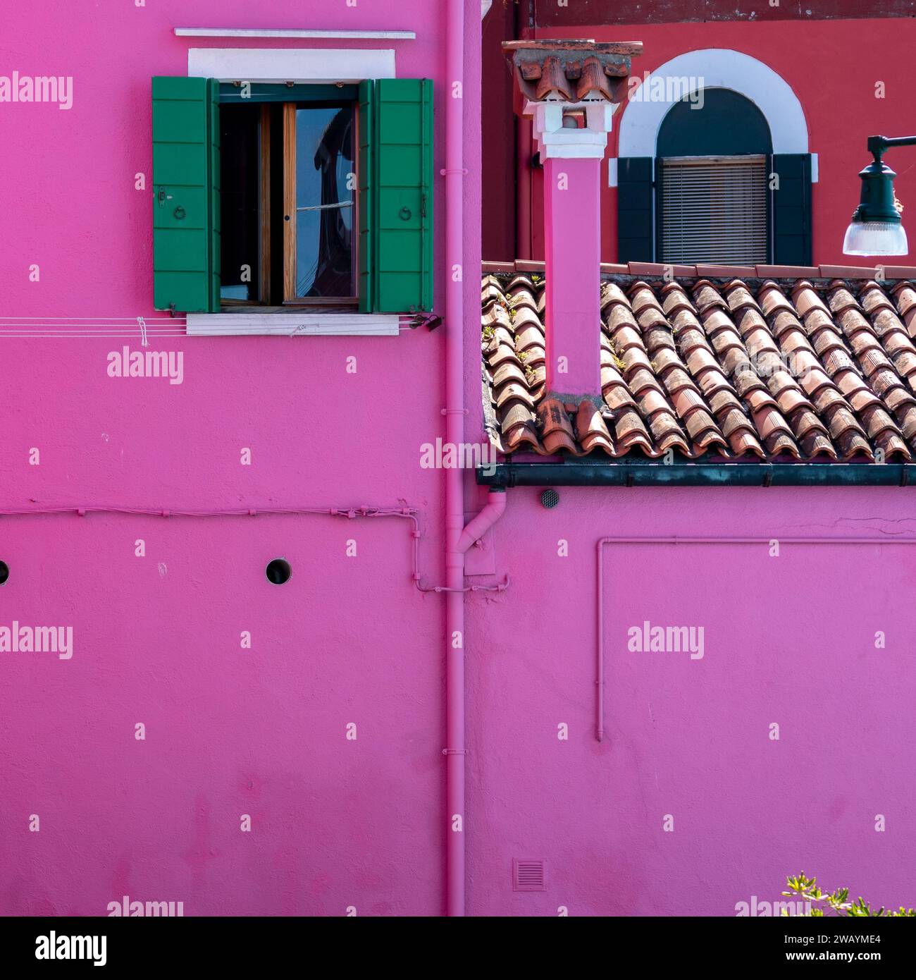 A vibrant pink building with green shutters and an open window sits atop a traditional brown roof Stock Photo