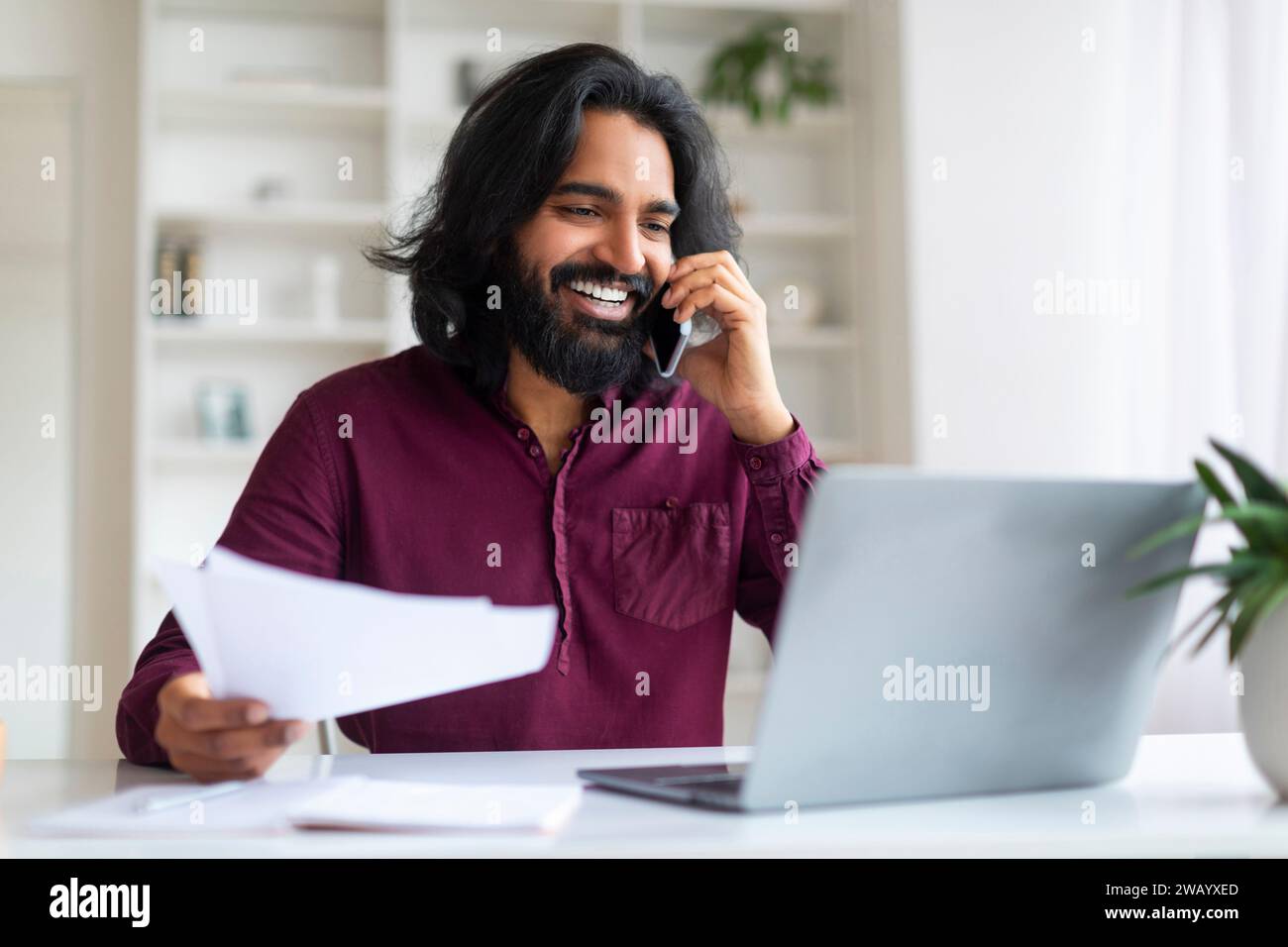 Indian man discussing work on phone while reviewing documents and using laptop Stock Photo