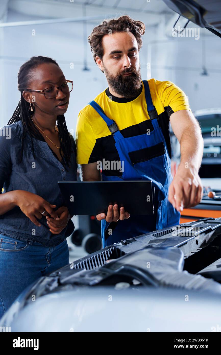 Mechanic in car service uses laptop to calculate invoice after fixing broken automobile distributor. Certified auto repair shop worker uses device to inform woman of final costs after mending vehicle Stock Photo