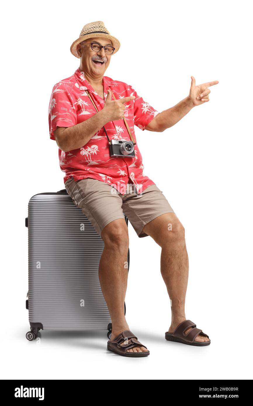 Excited male traveler sitting on a suitcase and pointing isolated on white background Stock Photo