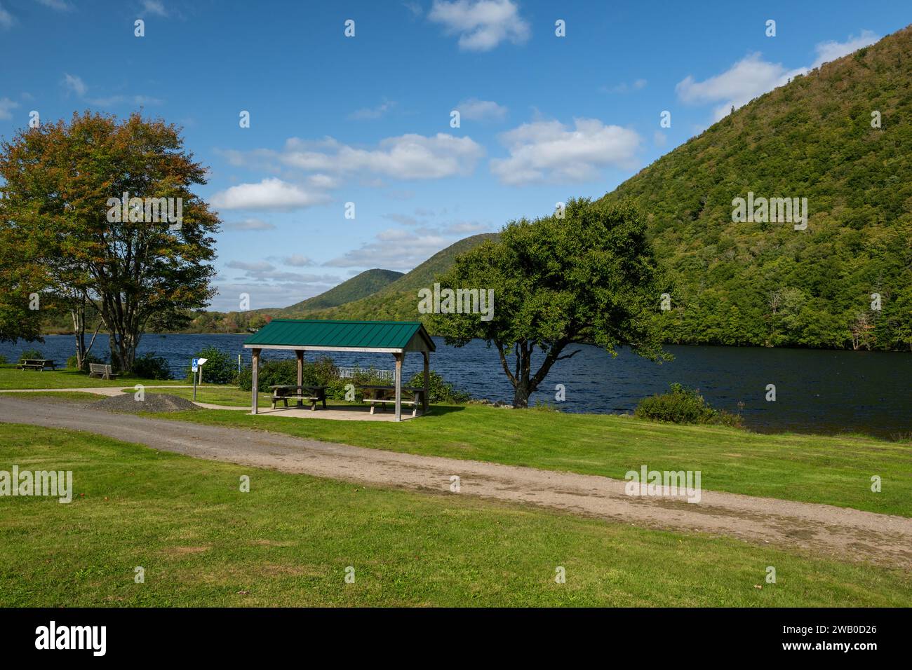 Picnic tables with a sheltered metal cover. The park campsite is near a river with tree-covered hills, lush green grass, and riverbanks. Stock Photo