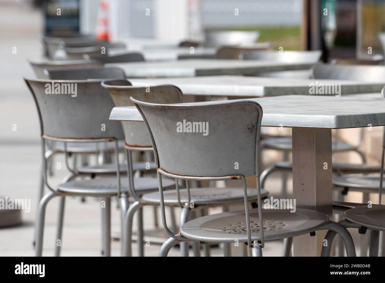 A row of multiple empty grey metal square tables and chairs outside on the sidewalk of a cafe or bakery. The chairs have round backs and are empty. Th Stock Photo