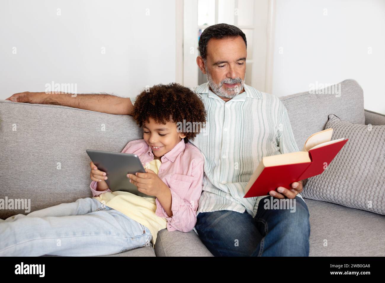 Senior Man Reading Book While Grandson Using Digital Tablet Indoor Stock Photo