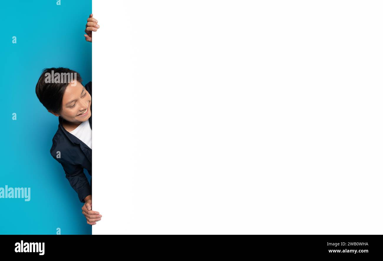 Asian man peeks playfully around a blank white vertical banner with a big smile Stock Photo