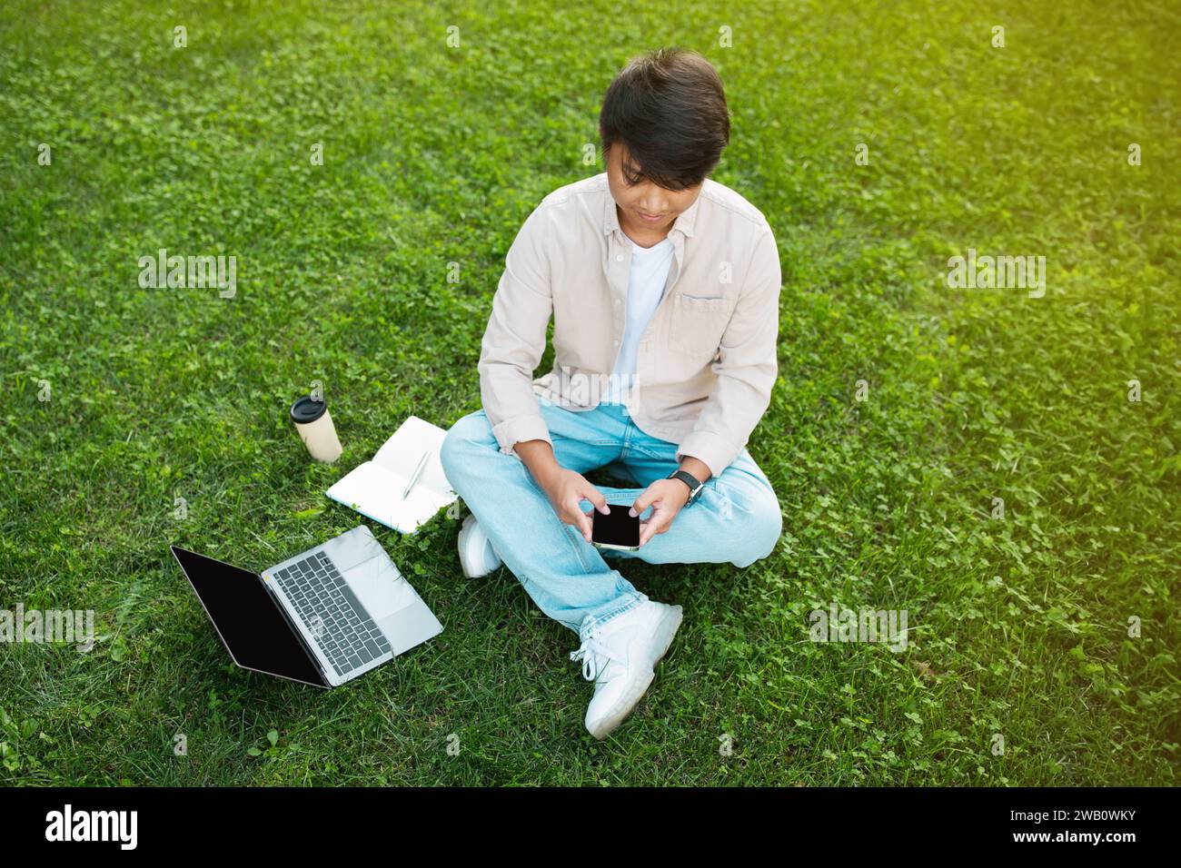 Asian man sitting on grass while using phone and laptop Stock Photo
