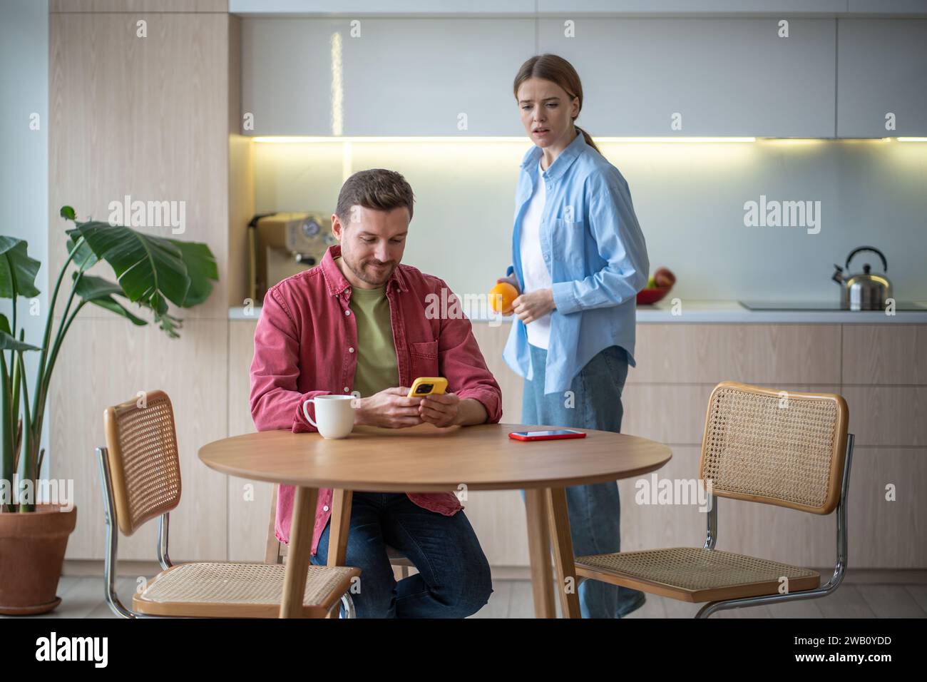 Jealous woman peeping at man smartphone on kitchen at home.  Stock Photo