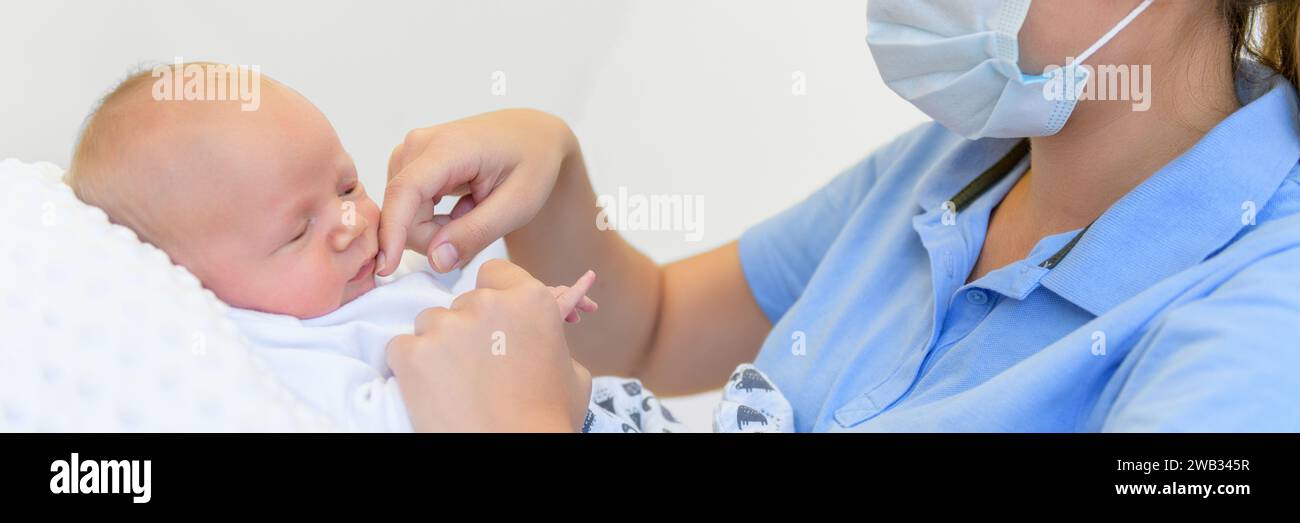 Midwife wearing face mask doing medical check on a newborn baby boy. Mother stroking infants face. Bonding, face massage, reflexology. Stock Photo