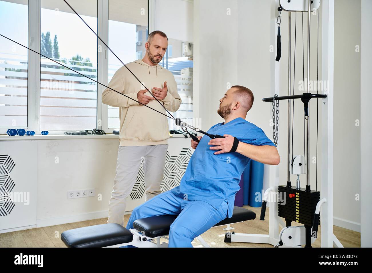 skilled rehabilitation specialist showing exercise on training machine to man in kinesiology center Stock Photo