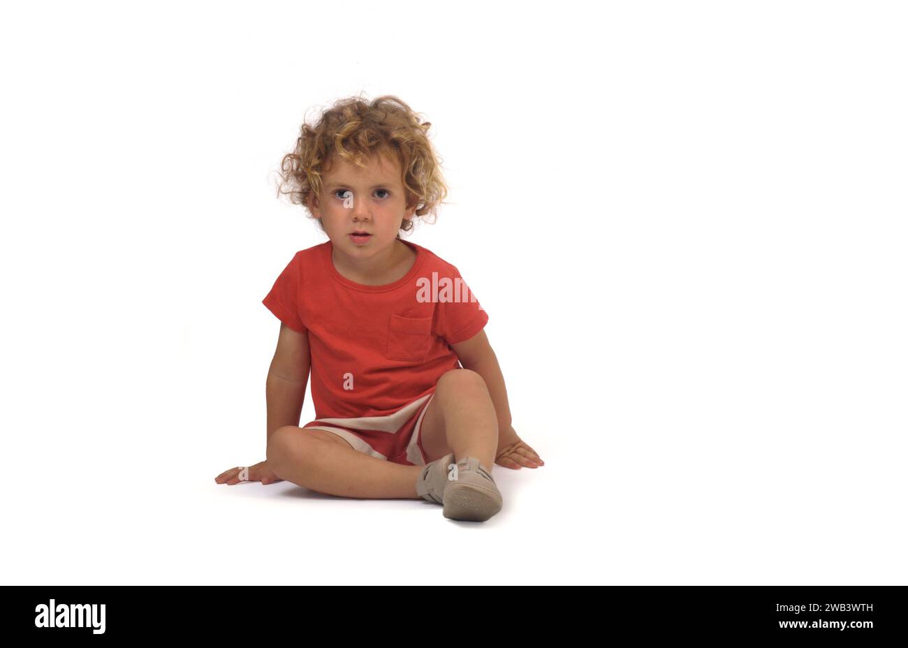 front view of a boy dressed in summer clothes sitting on group and looking at camera on white background (3 year old) Stock Photo