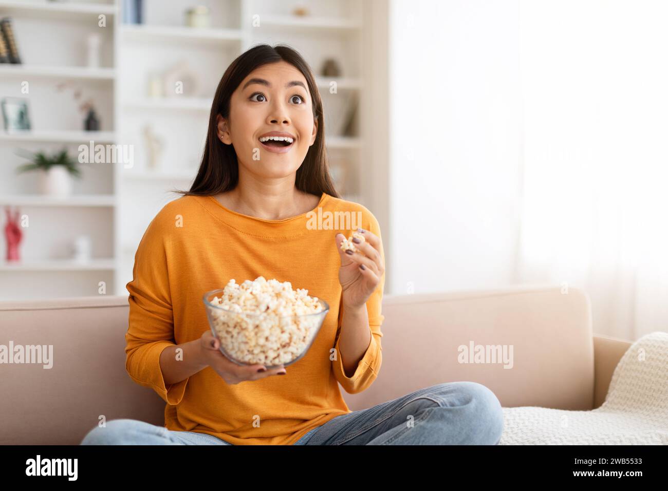 Amazed young chinese woman watching new TV show, eating popcorn Stock Photo