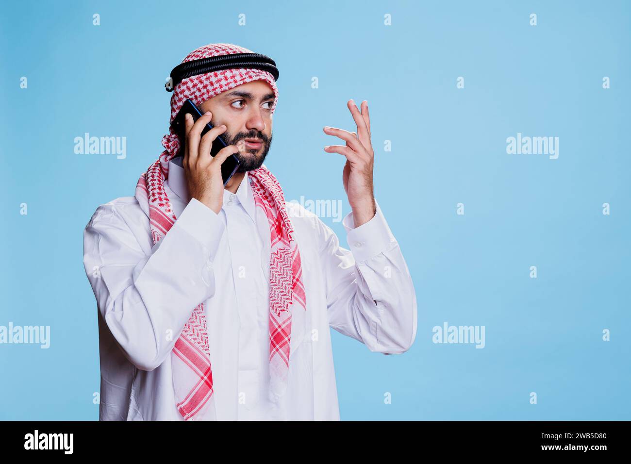 Muslim man wearing traditional clothes, speaking on smartphone and gesticulating with hands. Arab person dressed in white islamic thobe and ghutra talking on mobile phone Stock Photo