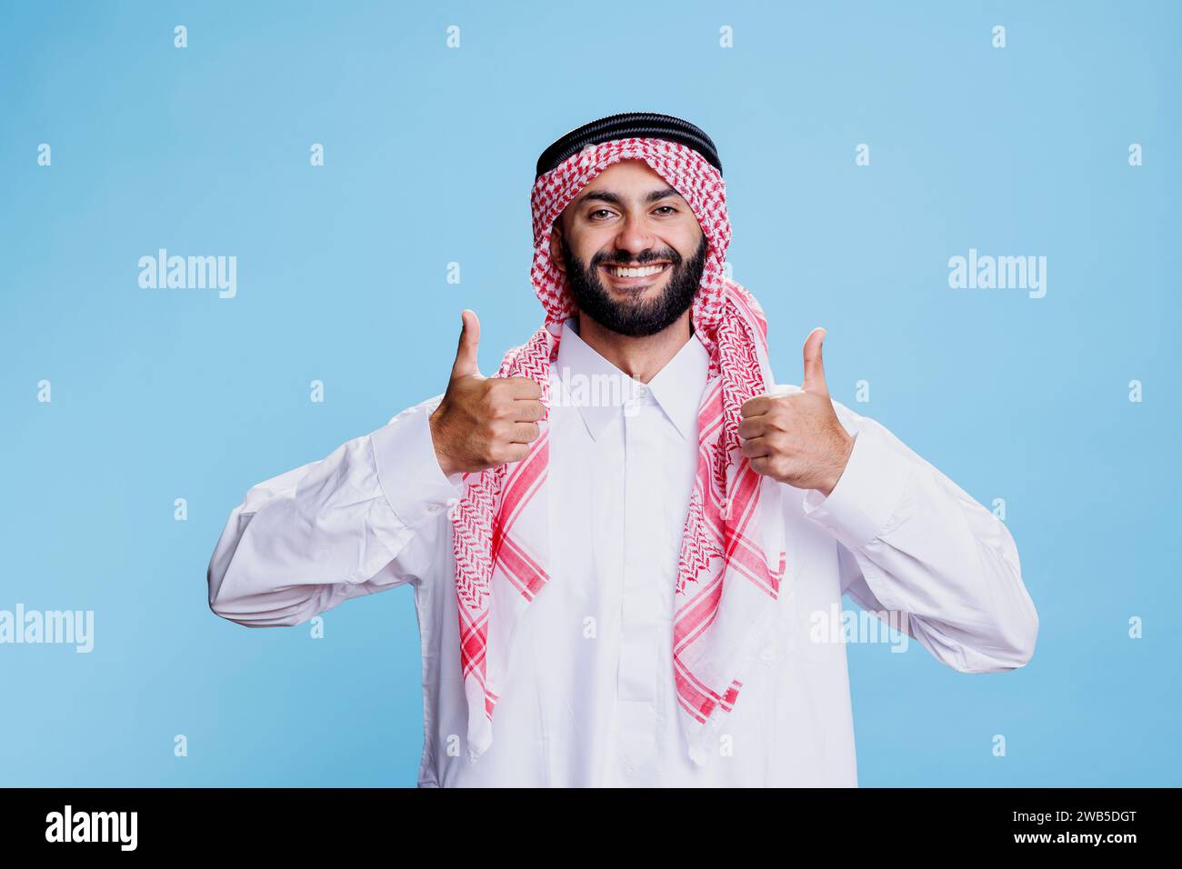Muslim man wearing traditional arab clothes standing with thumbs up gesture symbolizing agreement and positivity portrait. Cheerful arab dressed in thobe and headscarf showing approve sign Stock Photo