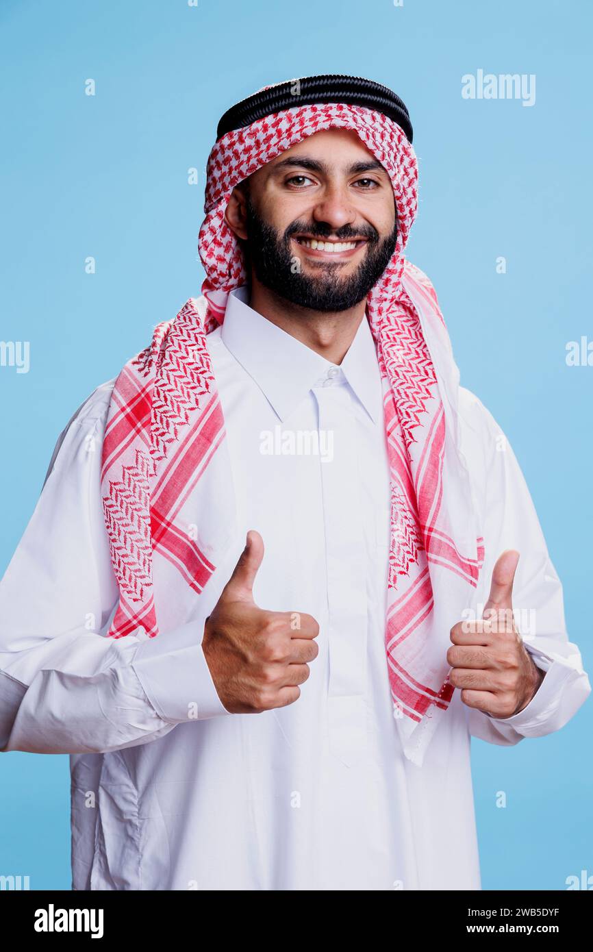 Cheerful muslim man wearing white thobe and headdress showing thumbs up studio portrait. Smiling arab person posing with positive gesture, showcasing arab culture and looking at camera Stock Photo