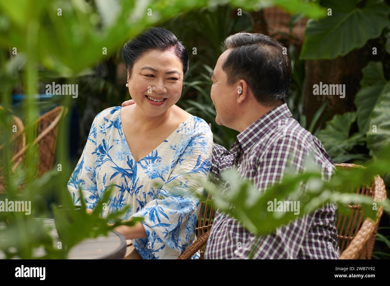 Happy senior Vietnamese man and woman sitting on bench in park and looking at each other Stock Photo