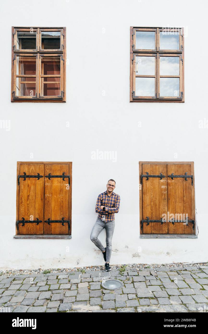 Young businessman in shirt leans his back against the wall of a white house in the historic centre of Sighisoara Stock Photo