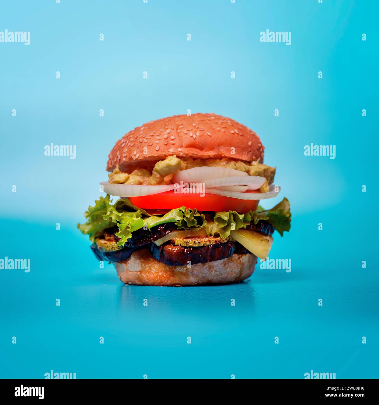 A freshly-made hamburger on a blue background, with lettuce, tomato, onion, and pickles. Stock Photo