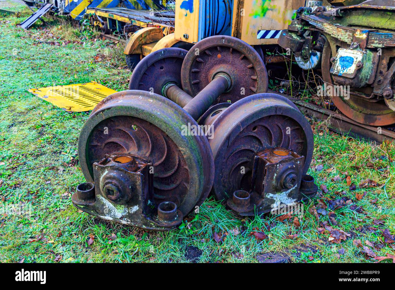 Two sets of train wheels stacked on green grass, parts of old freight railway, disused tracks in old station, remains of dilapidated carriages in back Stock Photo