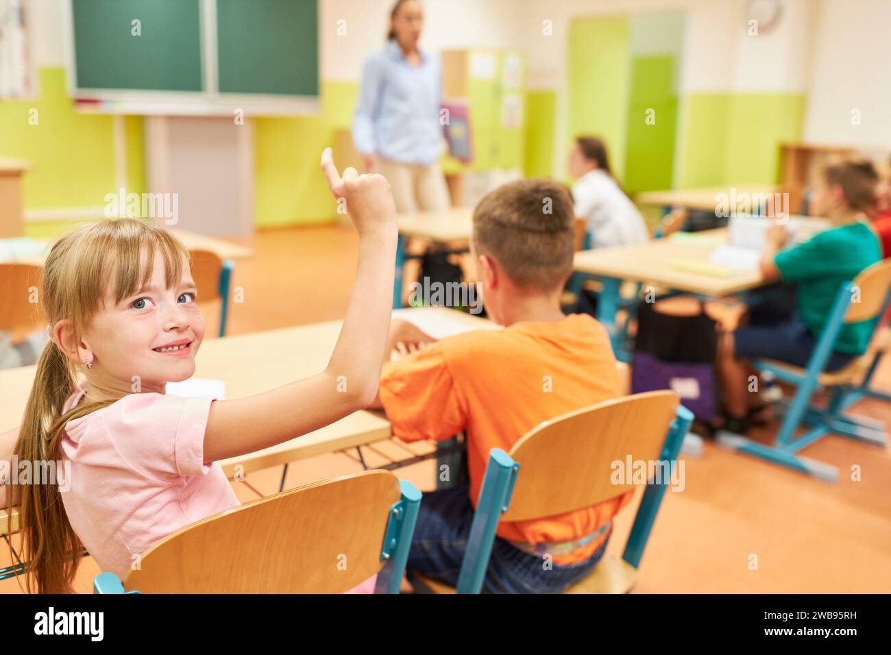 Smiling schoolgirl raising hand during QNA session while sitting with friend in classroom Stock Photo