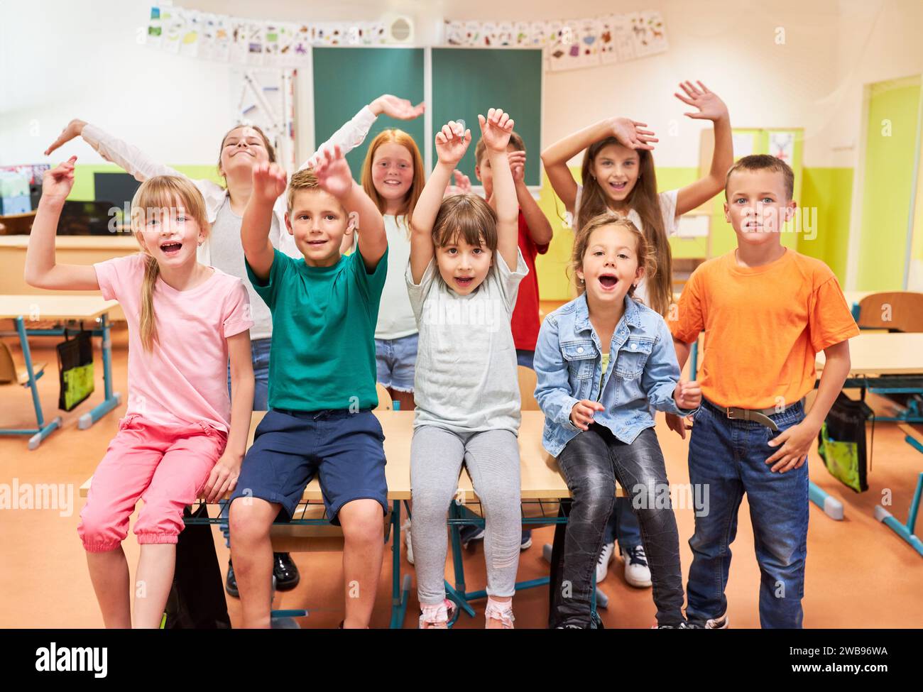 Portrait of happy elementary students sitting with arms raised on bench in class at school Stock Photo