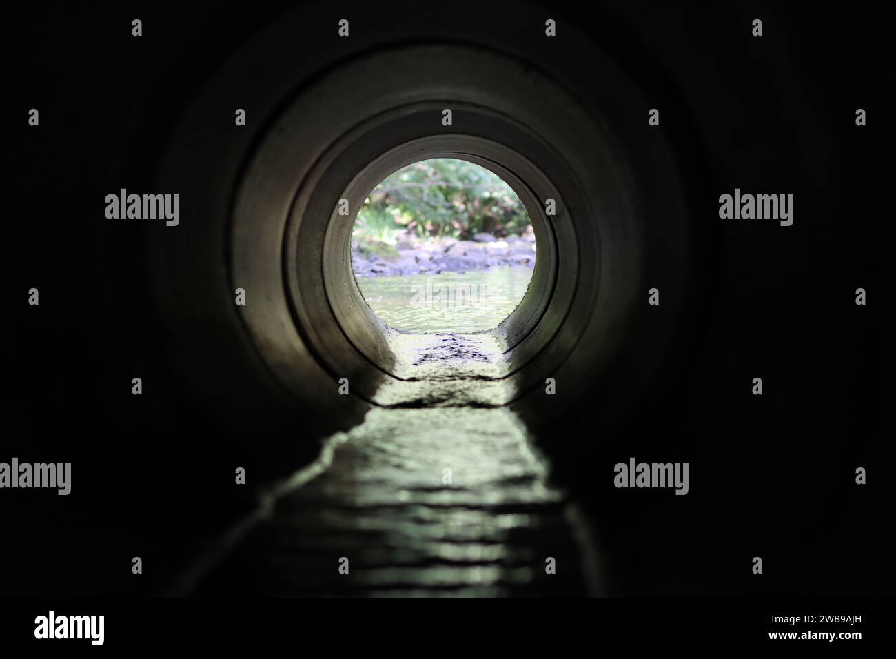 View through a round concrete tunnel carrying a stream beneath a road Stock Photo