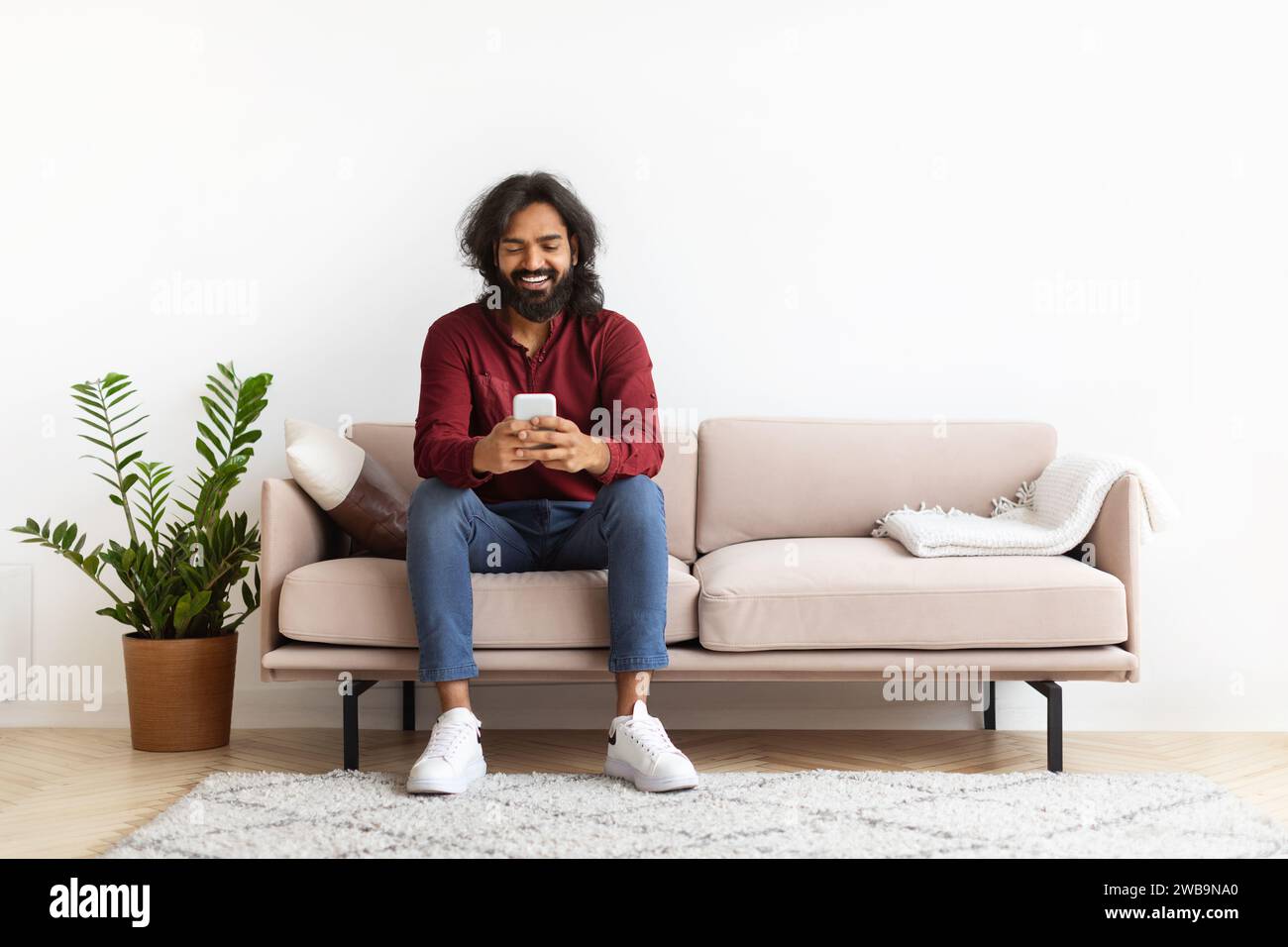 Cheerful young indian man sitting on couch, using phone Stock Photo
