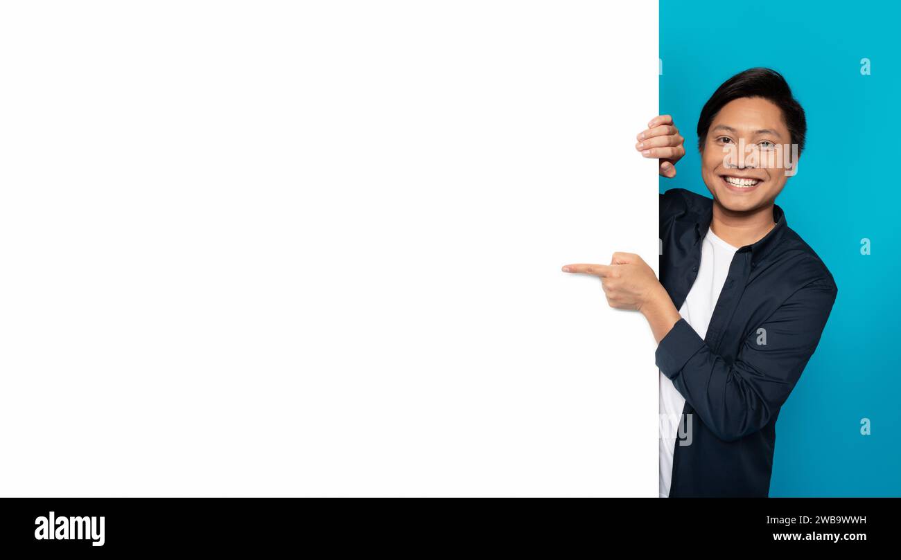 An Asian man with a cheerful smile is playfully peeking from behind a blank white vertical banner Stock Photo